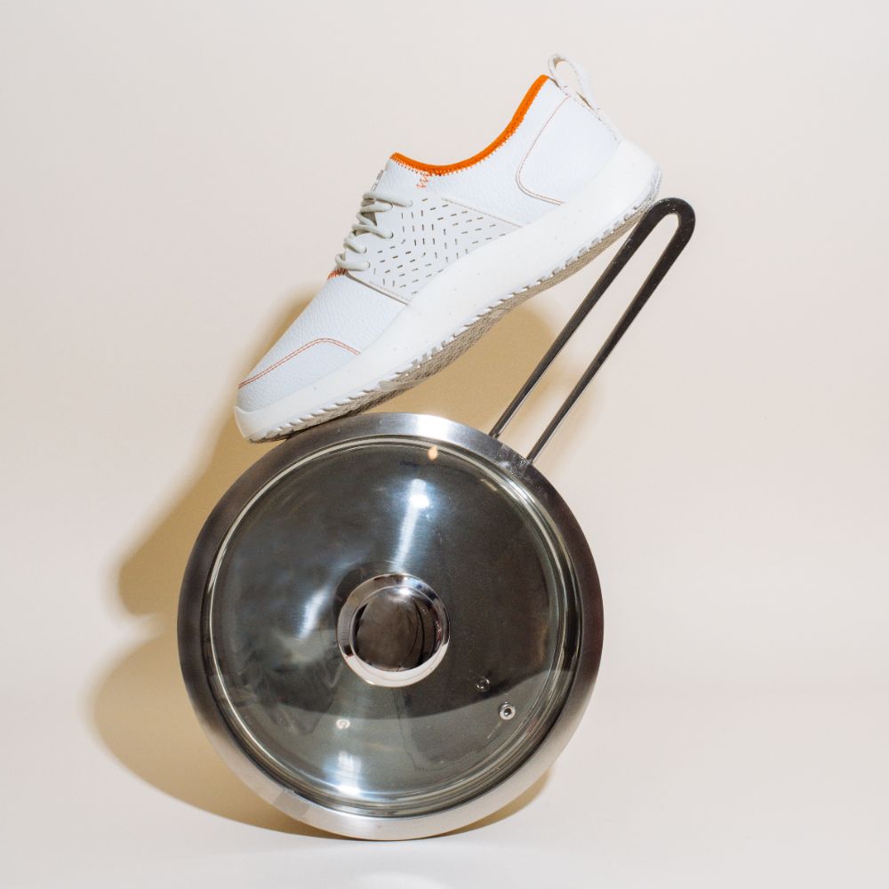 A Snibbs Men's Spacecloud Premium - Custard sneaker with crisp white vegan leather and orange accents balances on the handle of a glossy metal pot lid, its modern design standing out against the plain beige background.