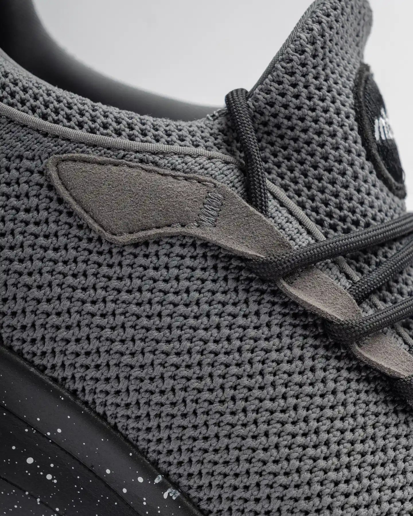 A detailed view of the Snibbs Orbit - Charcoal (2023) showcases its lightweight, breathable gray knit upper, complemented by black laces and suede accents. The sneaker features a black midsole with white speckles and a slip-resistant design for dependable