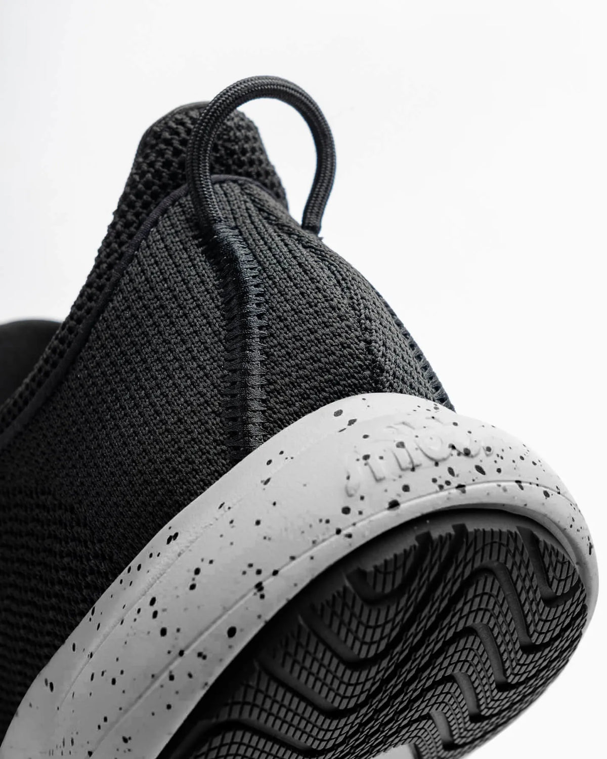 Close-up of the heel and sole of the Snibbs Orbit - Salt and Pepper, a black athletic shoe featuring a breathable knit upper and a white, speckled midsole, complemented by a textured black outsole for added style—photographed against a white background.
