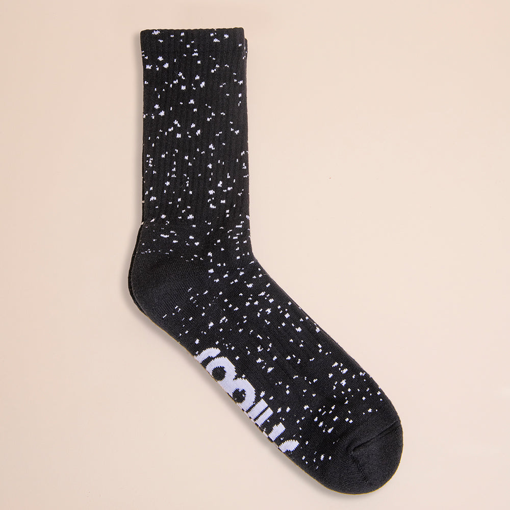 A Snibbs Crew Sock - Core Black with white speckles lies on beige, showing “rolling” partly visible on its sole.