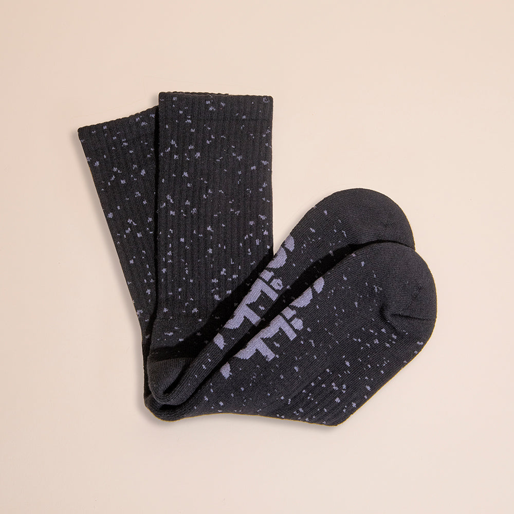 A pair of Snibbs Crew Socks - Core Eclipse, black with white speckles and text, seamless toe, displayed on a beige background.