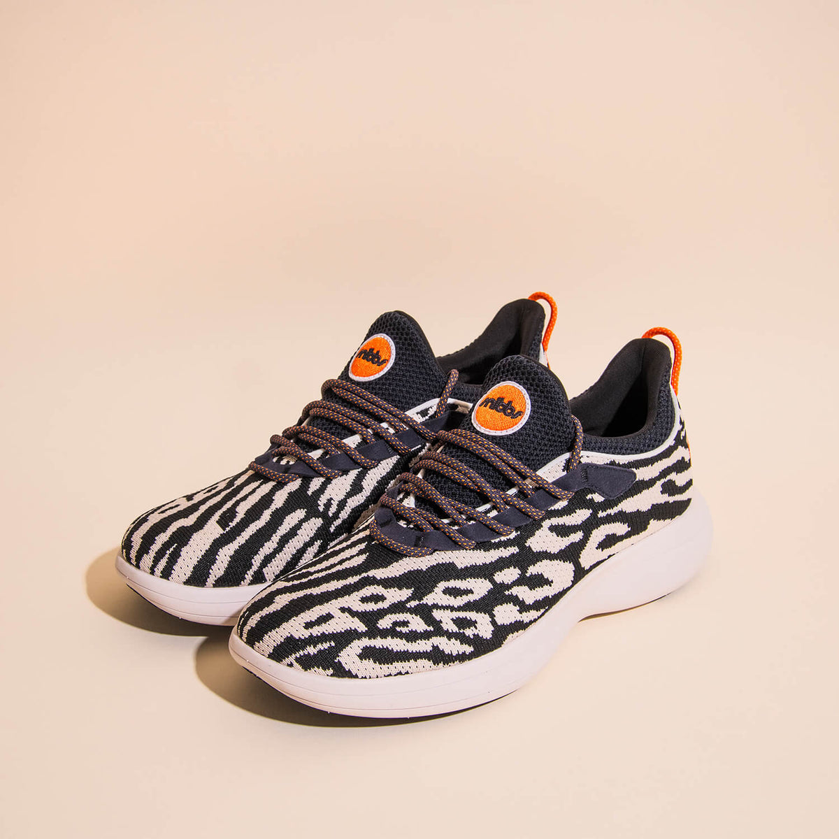 Snibbs Orbit - Snow Leopard features a lightweight, breathable knit upper with a bold black and white snow leopard print pattern, complemented by comfortable white soles and vibrant orange accents on the tongue and heel. These slip-resistant sneakers are 