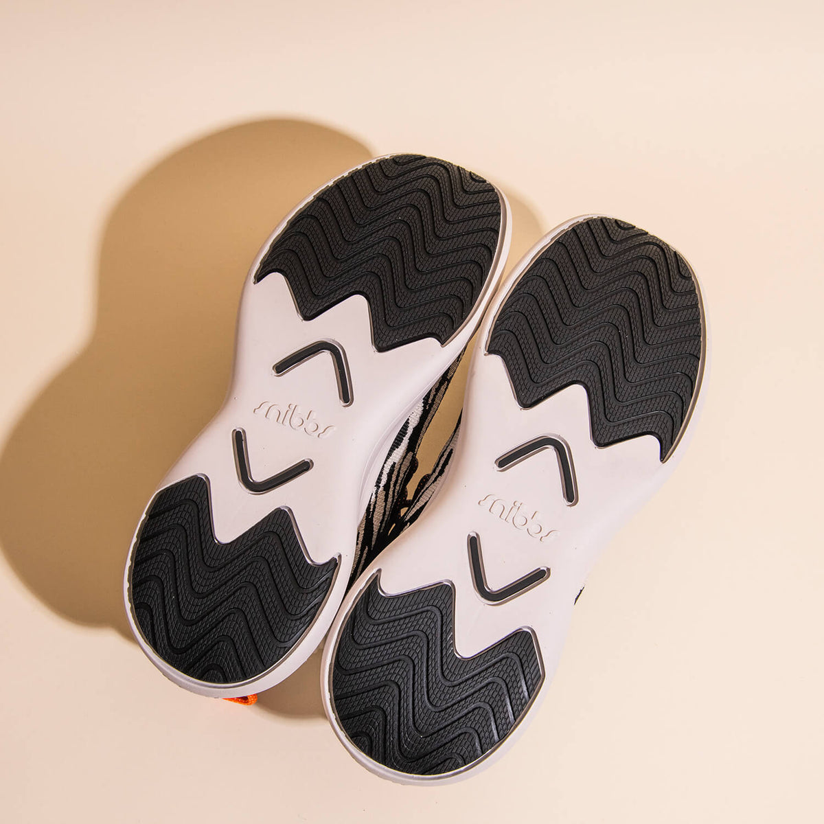 A pair of lightweight Snibbs Orbit - Snow Leopard sneakers shown from the bottom, displaying black and white outsoles with a wavy tread pattern and arrows designed for slip-resistant grip. The word "Snibbs" is embossed on each sole. The background is a so