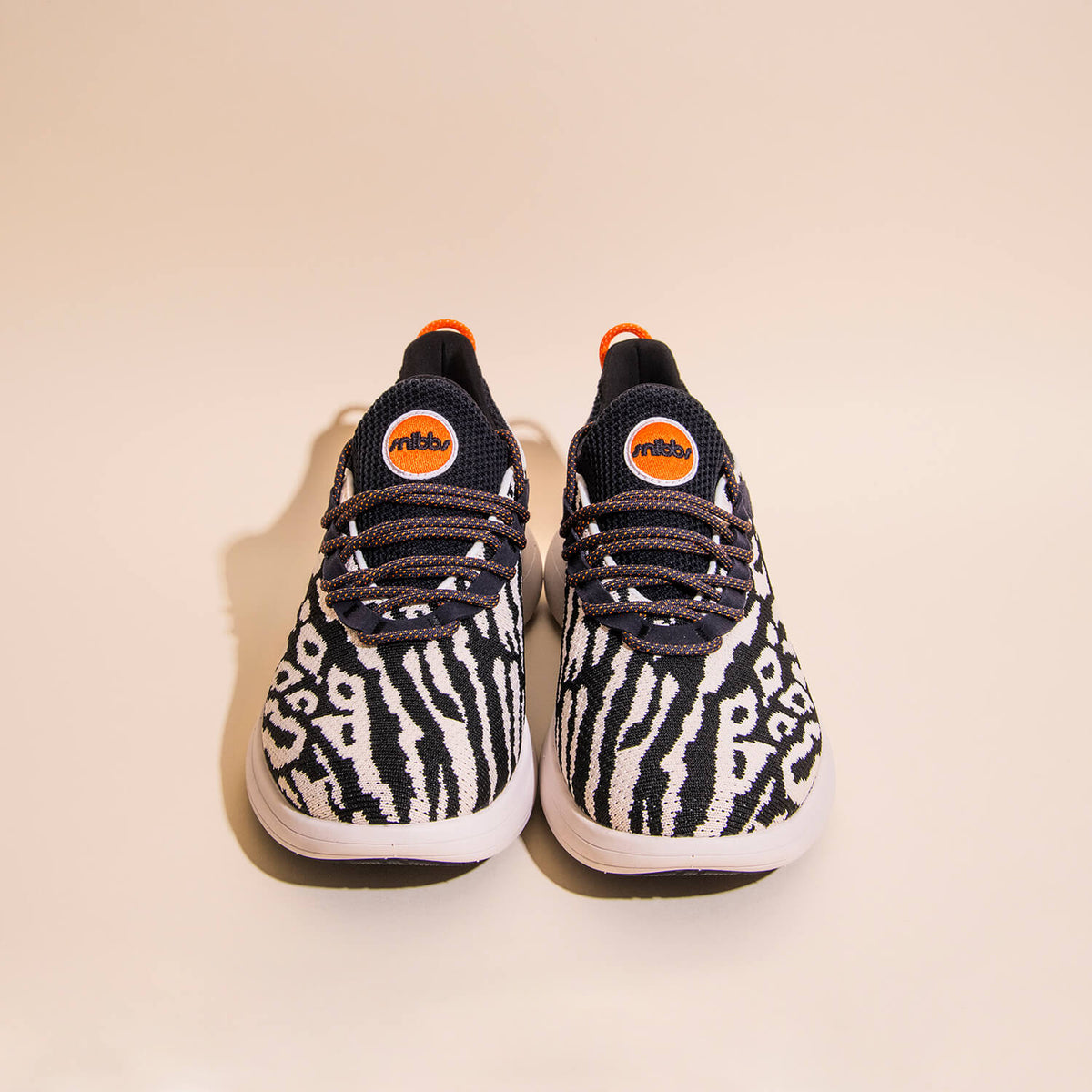 A pair of sneakers featuring a striking black and white snow leopard print, complemented by crisp white soles and an eye-catching orange accent on the tongue. Crafted with a lightweight, breathable knit upper for enhanced comfort, these sneakers are showc