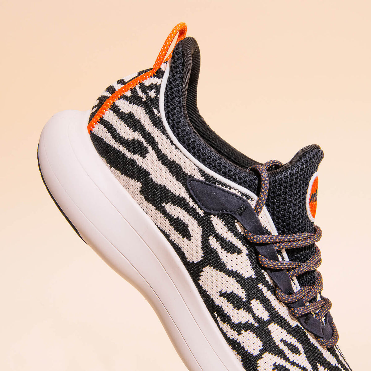 The Snibbs Orbit - Snow Leopard sneaker is showcased against a light peach background, featuring a black and white animal print pattern with orange accents, black laces, and a thick white sole. Designed for all-day comfort and style, this shoe offers a li