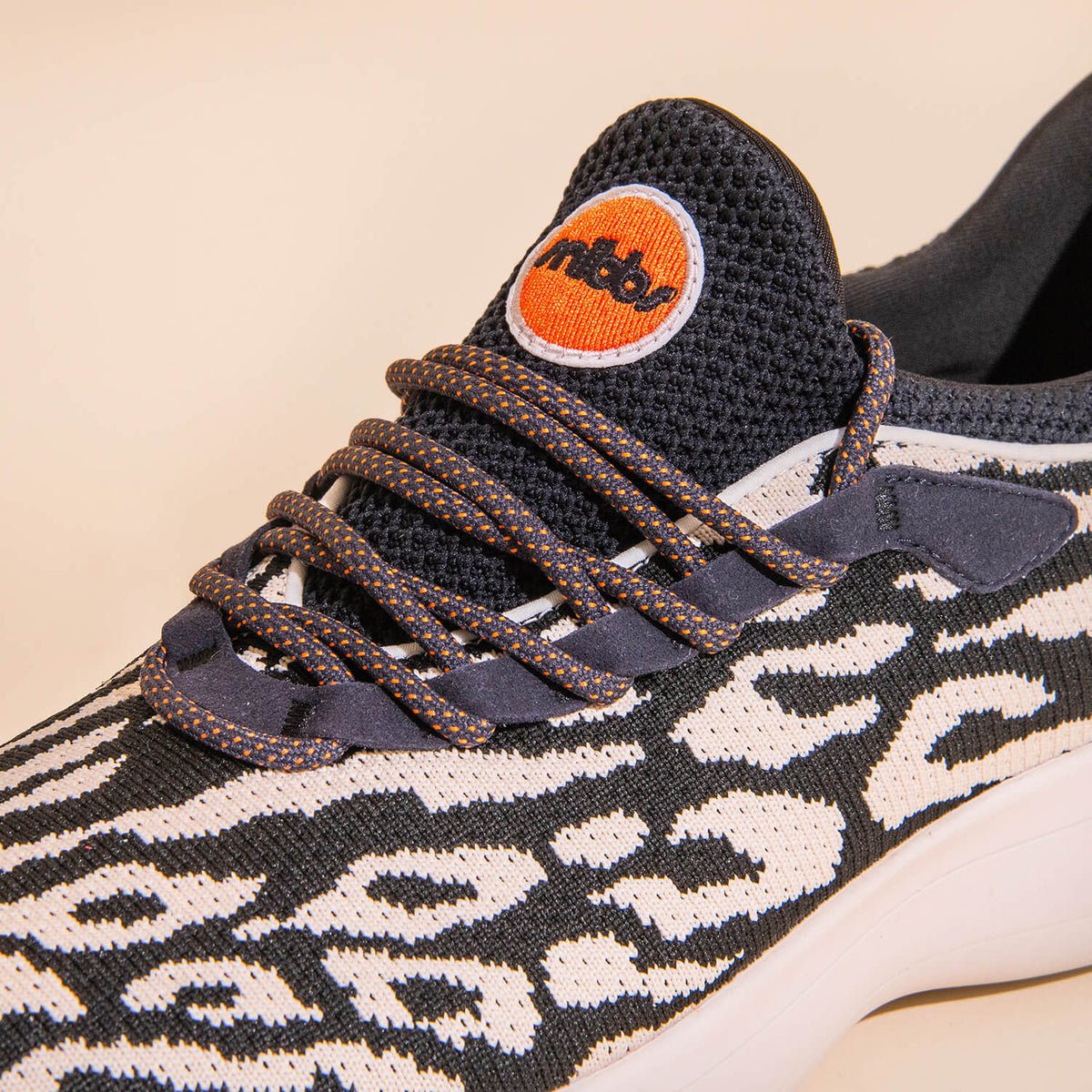 Close-up of the Snibbs Orbit - Snow Leopard sneaker, showcasing a black and white animal print pattern, dark laces, and an orange circular logo on the black mesh tongue. Designed with a breathable knit upper for enhanced comfort, this shoe also features a