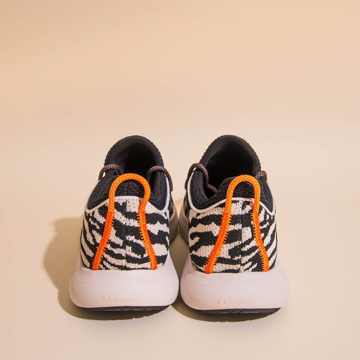 A pair of Snibbs Orbit - Snow Leopard sneakers, featuring a black and white snow leopard pattern on a breathable knit upper, white soles, and bright orange pull tabs on the heel, shown from the back against a neutral background.