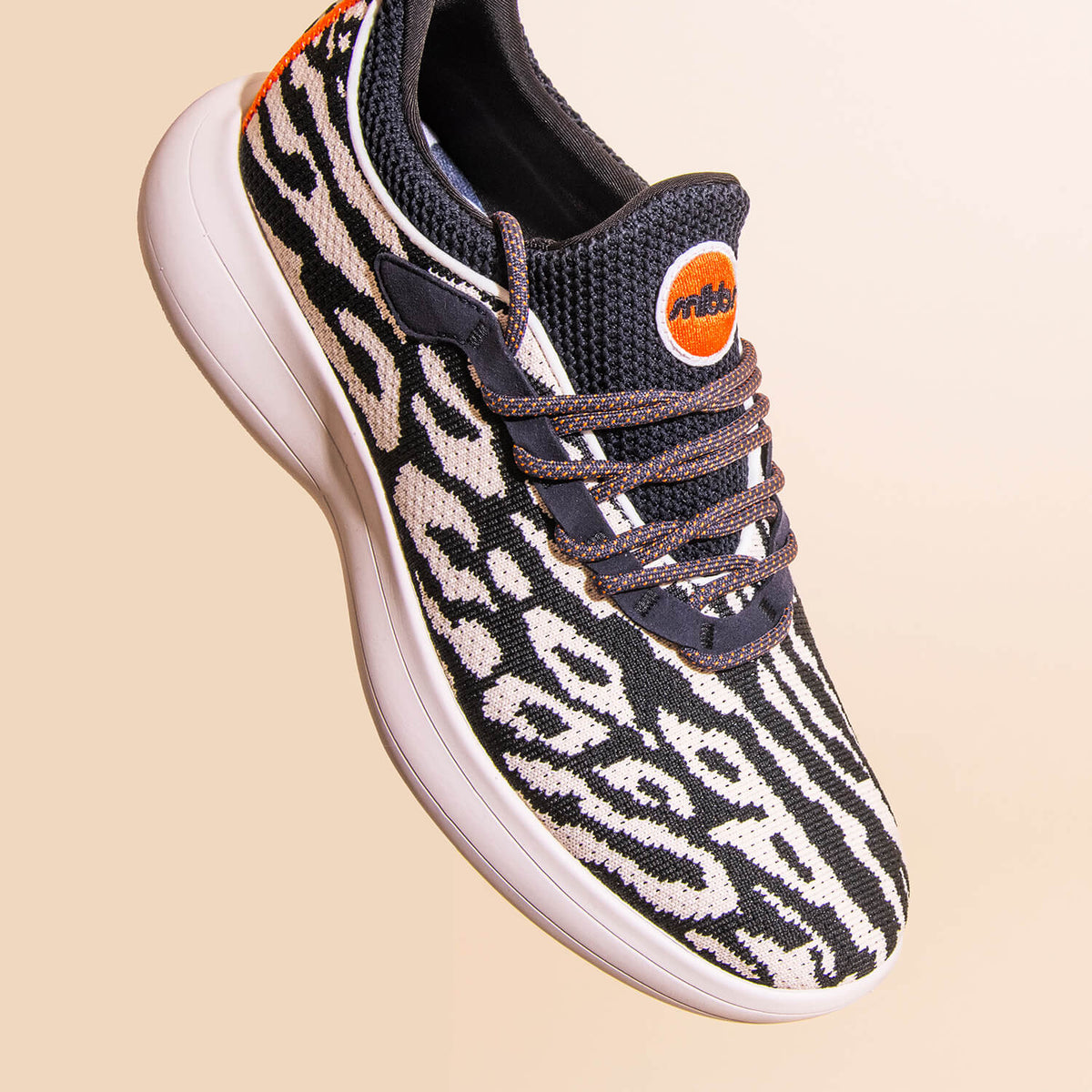 The Snibbs Orbit - Snow Leopard is a stylish sneaker featuring a black and white animal print pattern with bold orange accents and a white sole. Designed for all-day comfort, it boasts a lightweight, breathable knit upper and a slip-resistant design, show