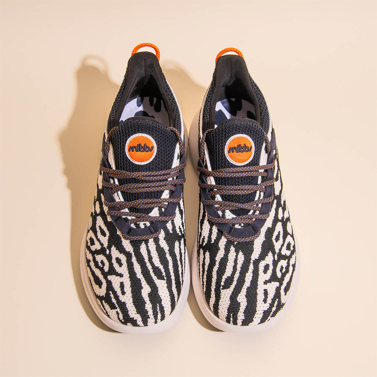 The Snibbs Orbit - Snow Leopard sneakers feature a striking black and white zebra-inspired pattern, round toes, thick white soles, and bold orange logo accents on the tongue. Designed with a breathable knit upper for enhanced comfort, they are photographe