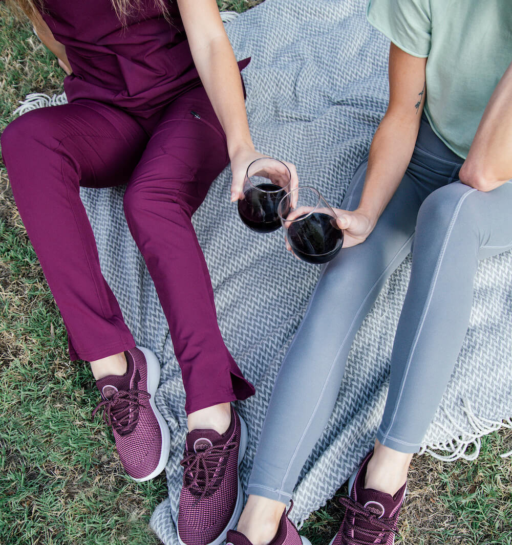 Two seated outdoors wear Snibbs Orbit slip-resistant shoes and athletic wear, holding red wine; only their lower bodies are visible.