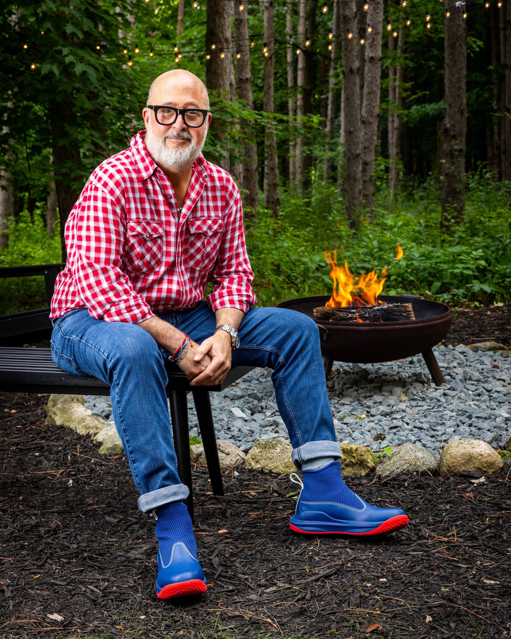 Andrew Zimmern sitting by a fire pit in a forest setting wearing a red checkered shirt, blue jeans, and blue rain boots.