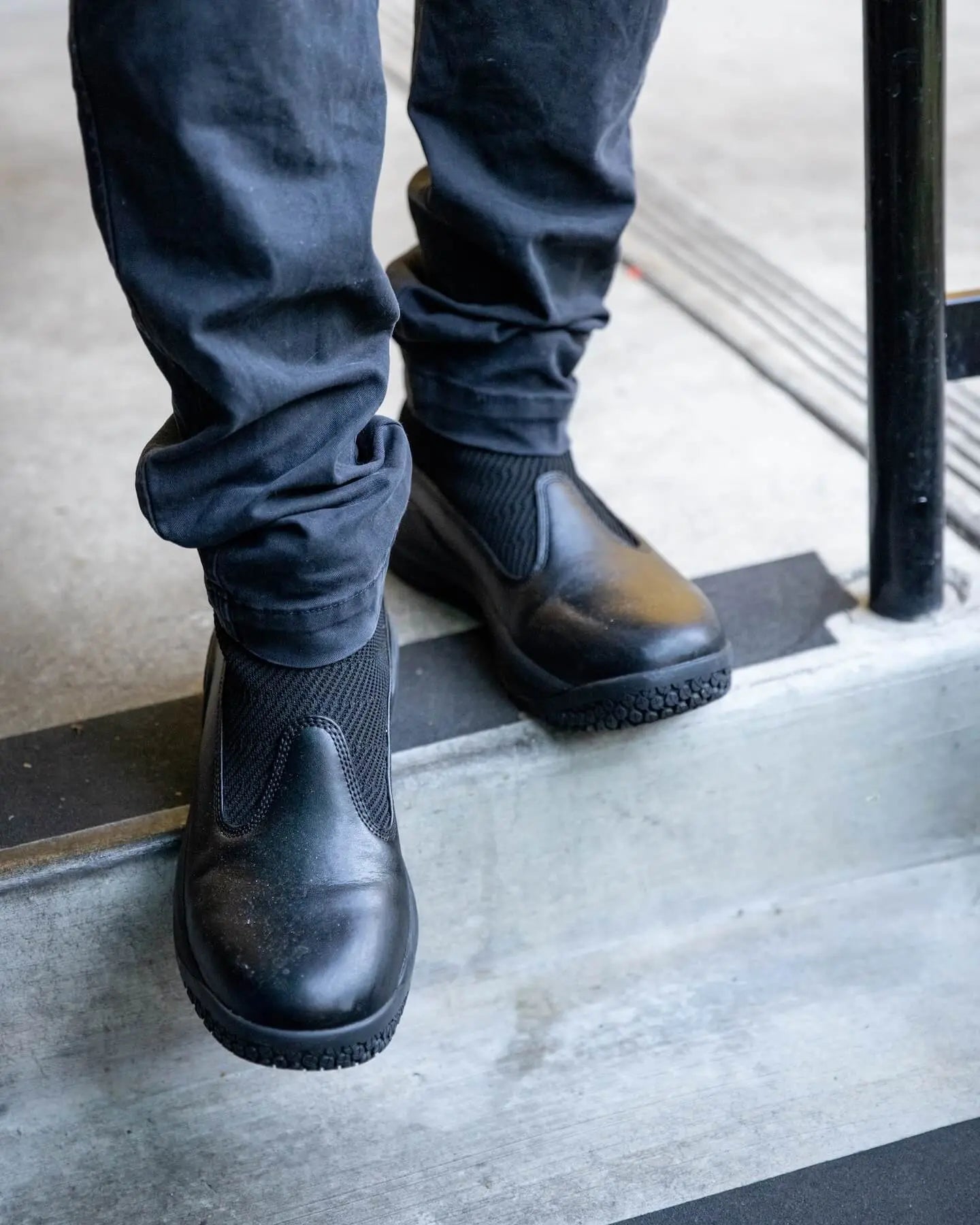 Someone wearing Snibbs Pro - Eclipse Black slip-ons with a premium knit upper and dark pants stands on a concrete step by a black rail.