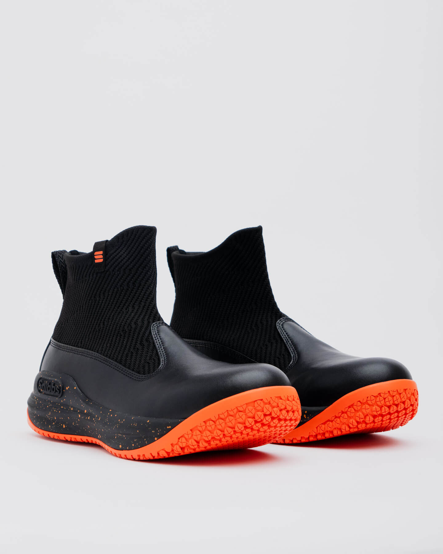 The Snibbs Pro - Tangerine: black slip-on ankle boots with textured orange soles, shown on a light gray background.