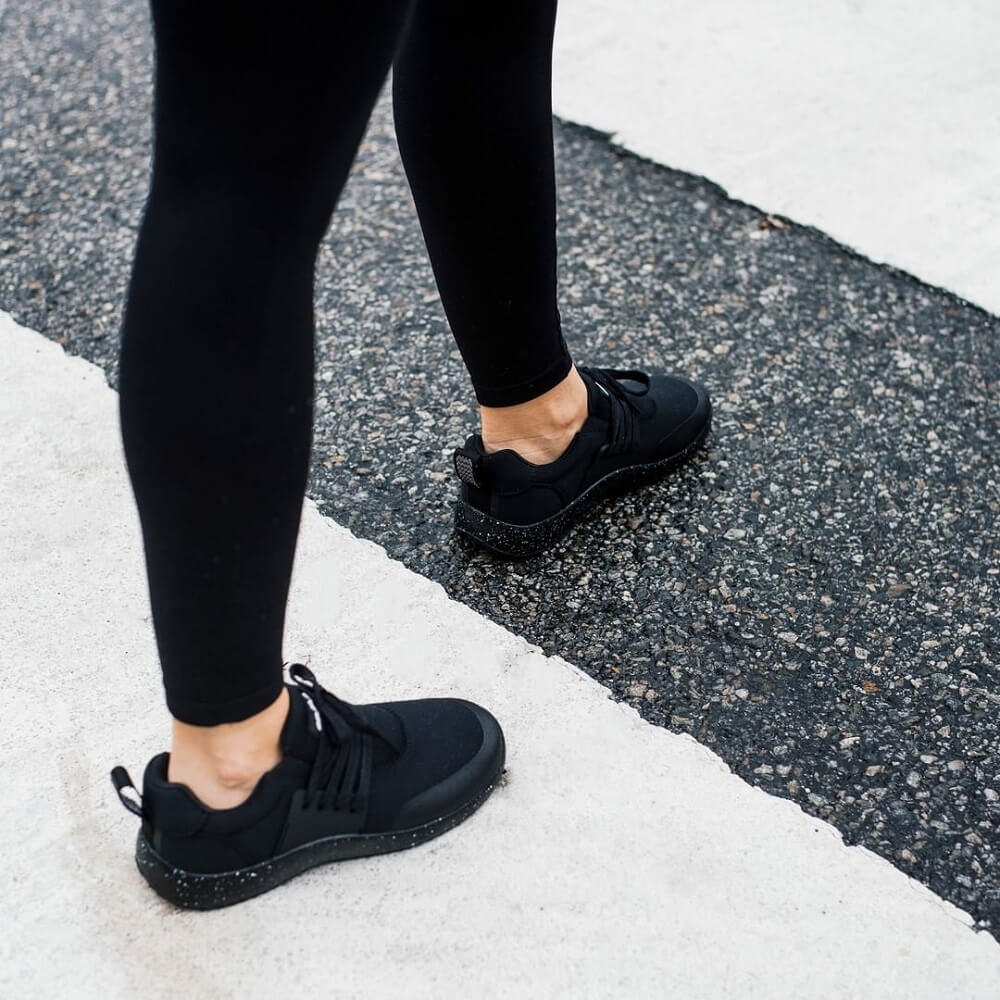 A person is walking on a paved surface with white and black sections, wearing black leggings and Snibbs Women's Spacecloud - Black Speckle sustainable, slip-resistant work shoes. Only their legs and feet are visible.