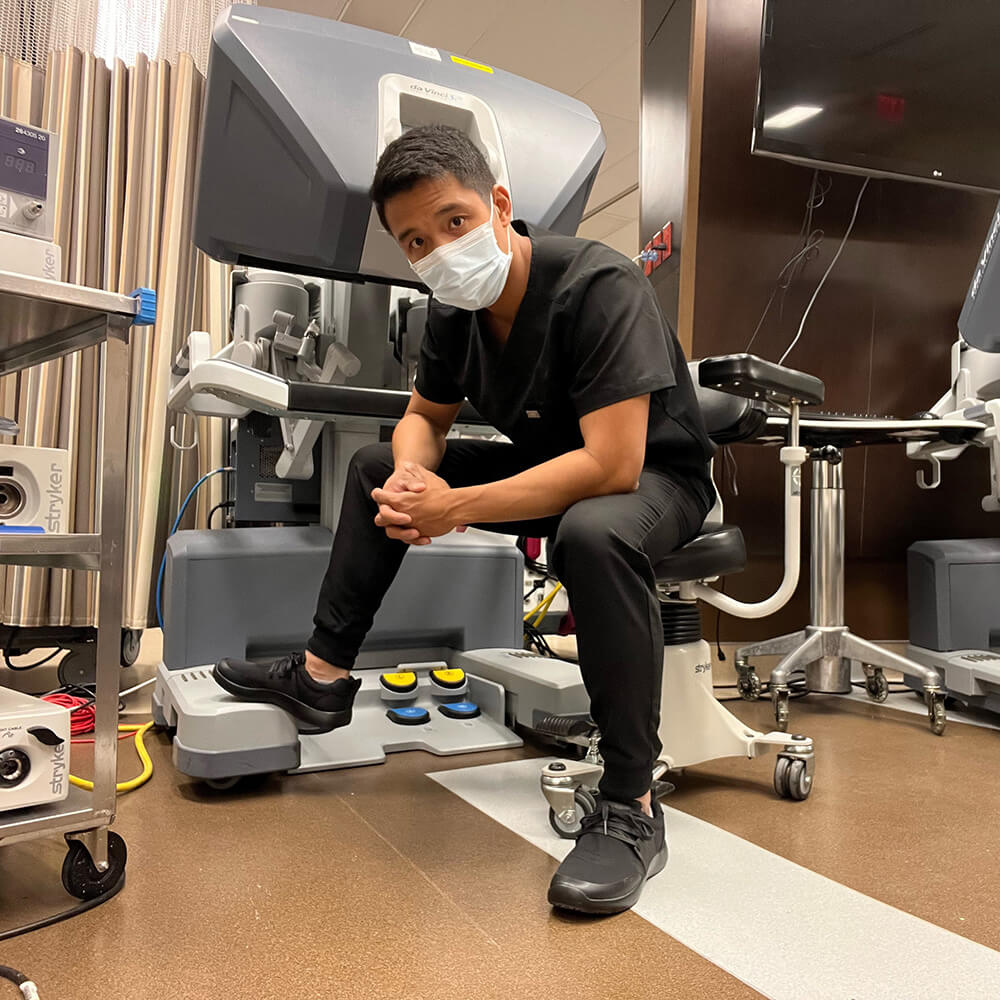 A person in black scrubs and a face mask, wearing Snibbs Men's Spacecloud - Eclipse Black slip-resistant work sneakers made from recycled materials, sits on a medical chair in front of advanced medical equipment in a clinical setting.