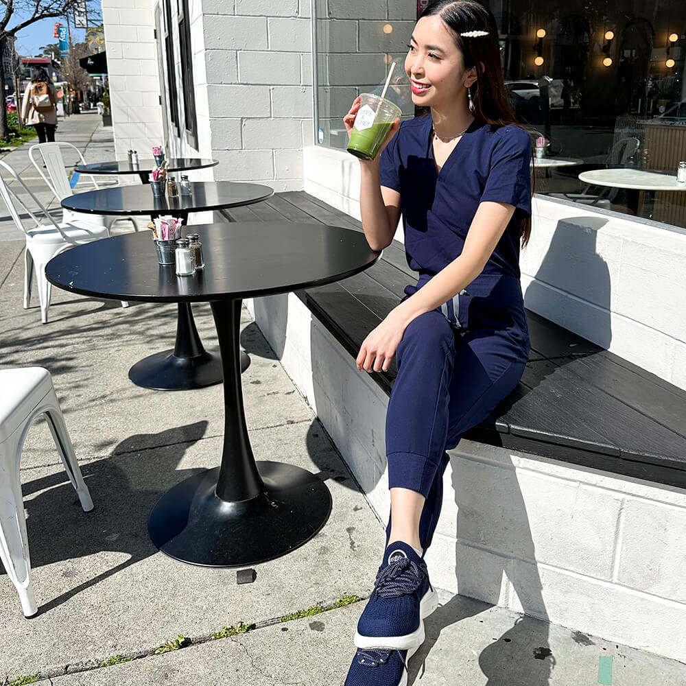 A woman in Snibbs Orbit - Maritime Blue slip-resistant shoes relaxes outdoors, enjoying a green drink on a sunny day.