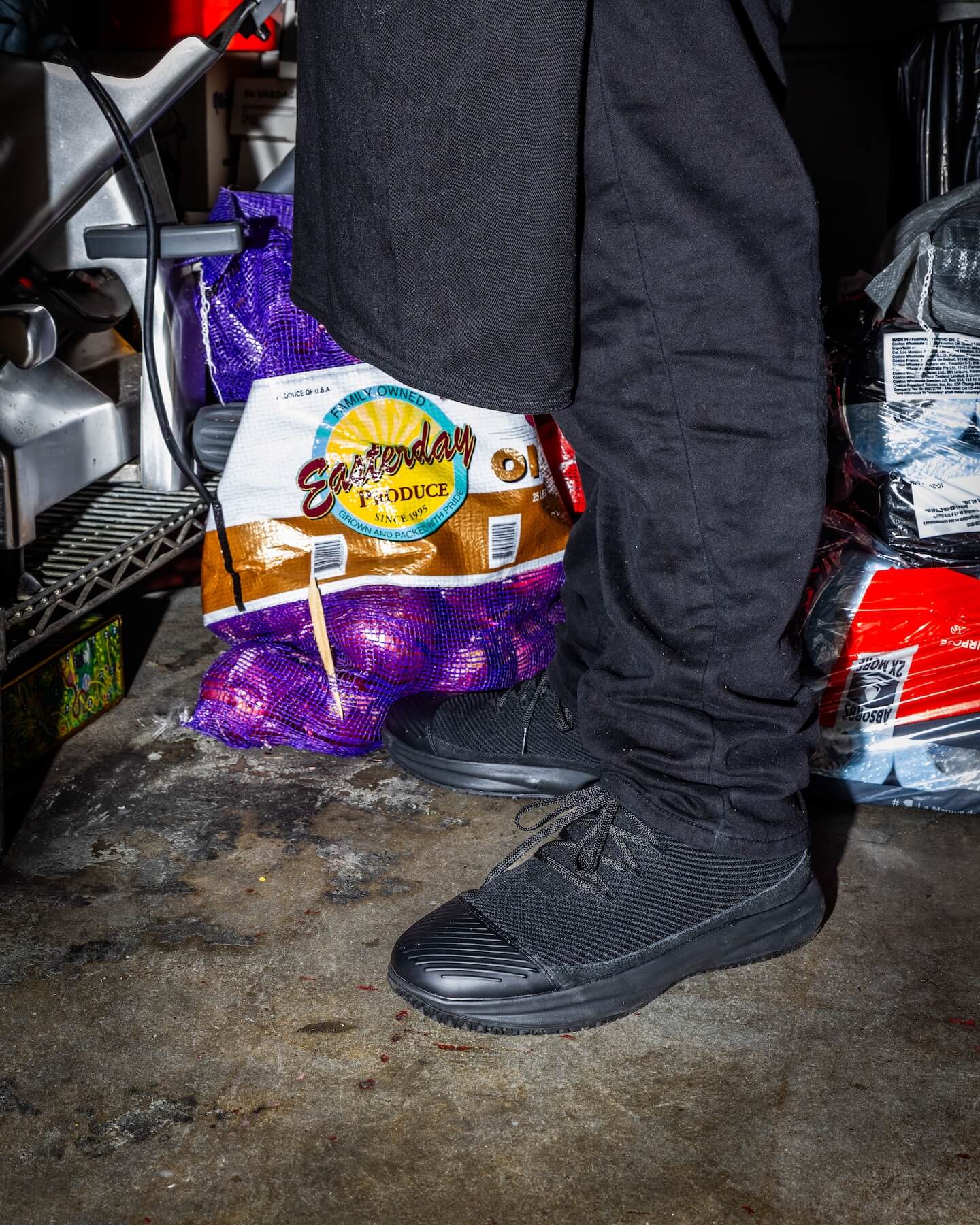 Person in Snibbs ROVR Mid - Climbing Ivy stands on a concrete floor near bags of produce, including a sack of red onions.
