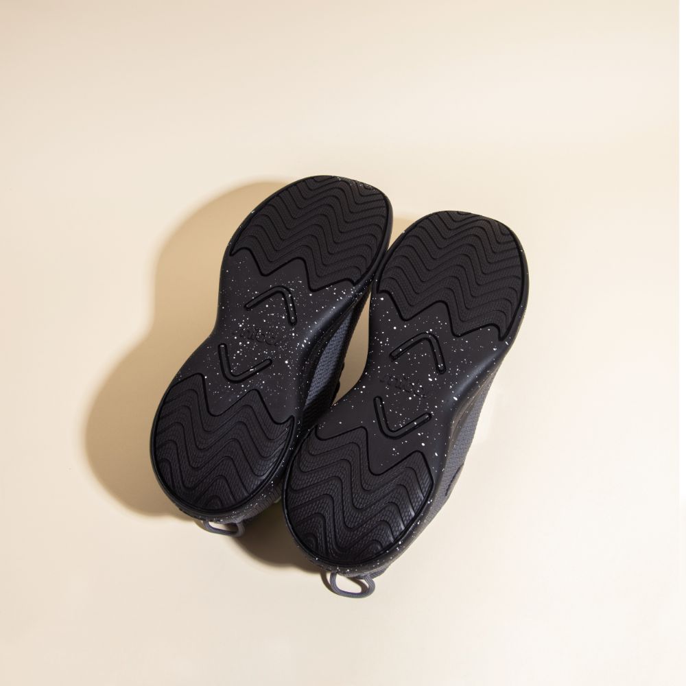 A pair of Snibbs Orbit - Charcoal shoes, shown from below with slip-resistant wavy tread and white specks on textured soles.