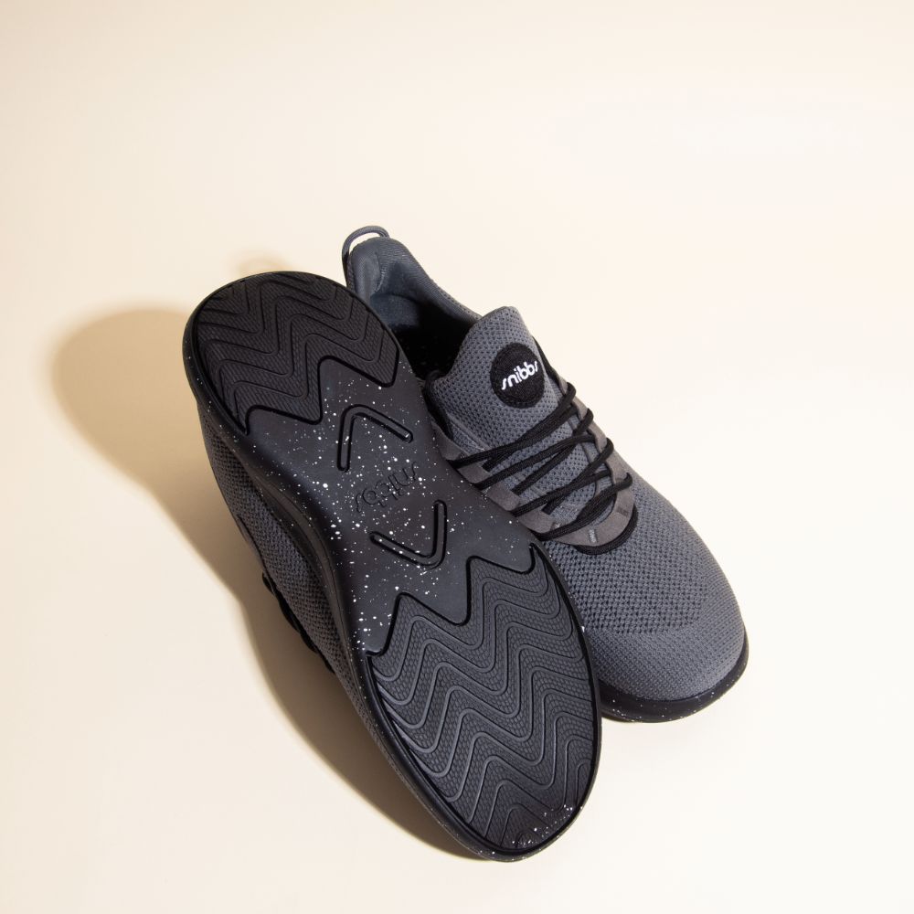 Orbit - Charcoal by Snibbs: gray athletic shoes with breathable knit uppers and black slip-resistant soles, one upright, one showing tread.