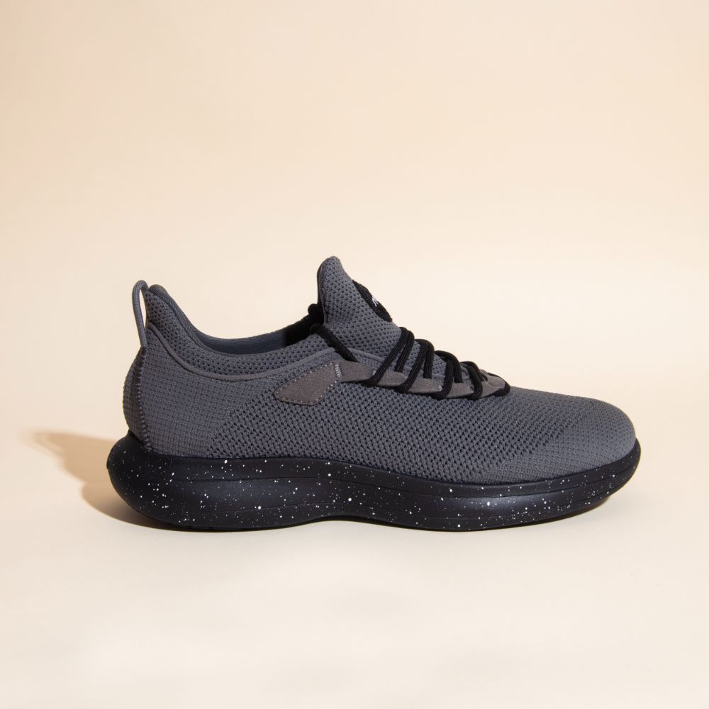 The Snibbs Orbit - Charcoal, a gray knit athletic shoe with black speckled sole, is shown on a beige background facing right.