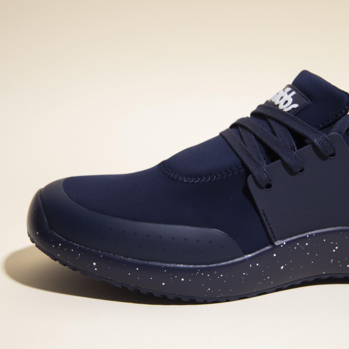 Close-up of the Snibbs Women's Spacecloud sneaker in Nebula Blue, featuring black laces, a smooth vegan upper, and a textured sole with white speckles. This stylish work sneaker is displayed against a light beige background.