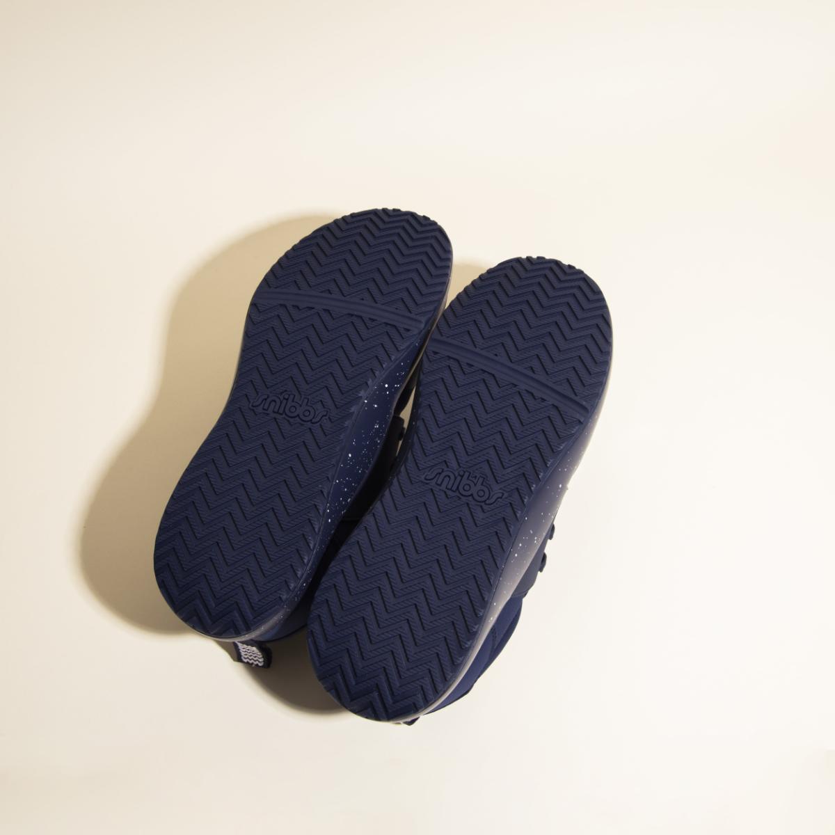 A pair of Snibbs Women's Spacecloud sneakers in Nebula Blue is shown from the bottom, featuring a herringbone tread pattern on the soles for slip-resistant grip, with "snibbs" embossed in the center of each sole.
