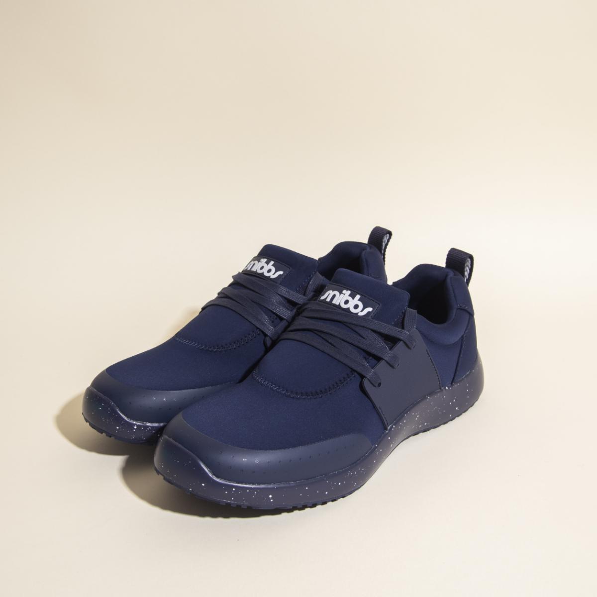 A pair of Snibbs Women's Spacecloud - Nebula Blue vegan sneakers, featuring black speckled, slip-resistant soles and snibbs branding on the tongue, displayed on a light beige surface.