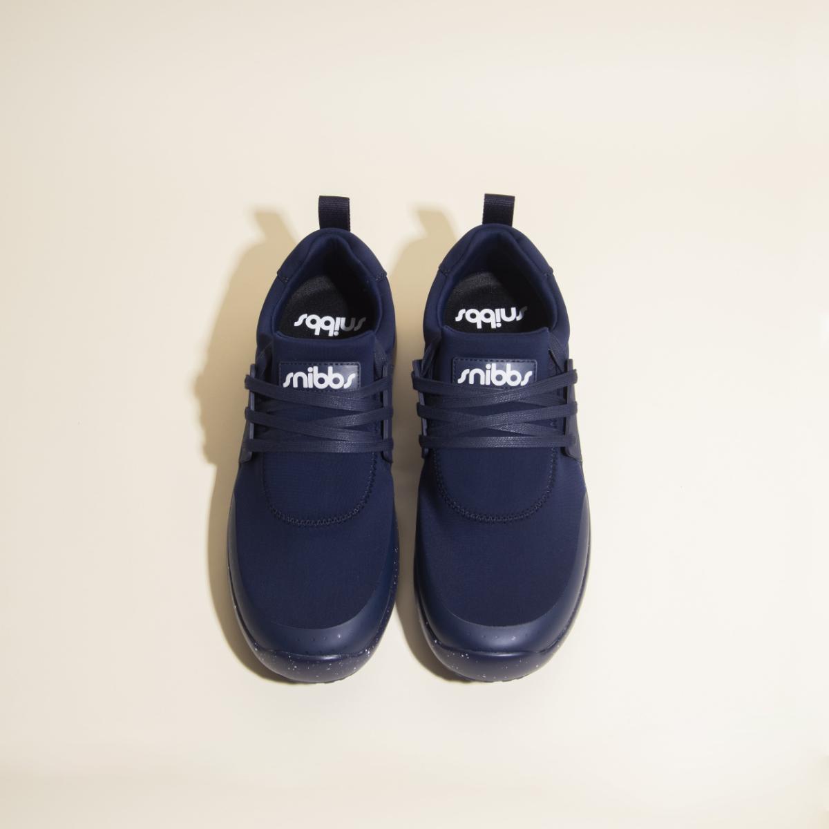A pair of Snibbs Women's Spacecloud sneakers in Nebula Blue, featuring white branding on the tongue, viewed from above on a light beige background. These slip-resistant shoes are thoughtfully designed to provide both comfort and style.